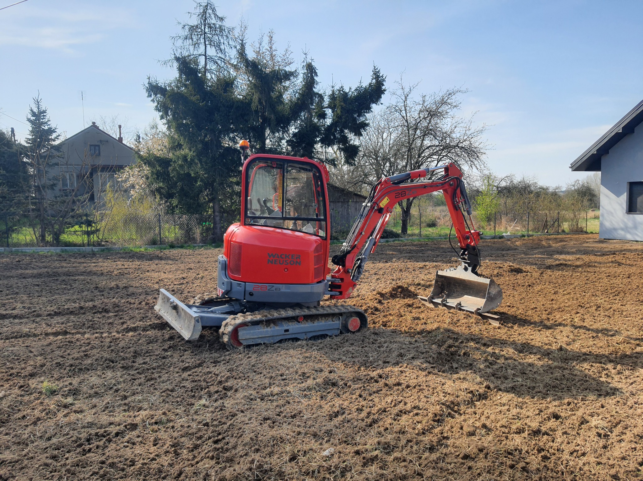 Czerwona mini koparka Wacker Neuson 28Z na wyrównanym, brązowym terenie. W tle domy i drzewa pod błękitnym niebem.