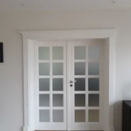 Białe, dwuskrzydłowe drzwi z matowymi szybami w ramkach, z ozdobną listwą górną, zamontowane w jasnym pomieszczeniu z parkietem.