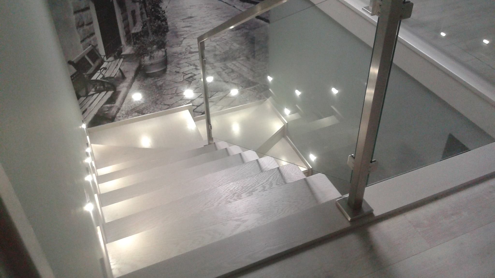 Nowoczesne schody z jasnego drewna z podświetleniem LED w stopniach i szklaną balustradą, widok z góry, na ścianie fototapeta z motywem uliczki.