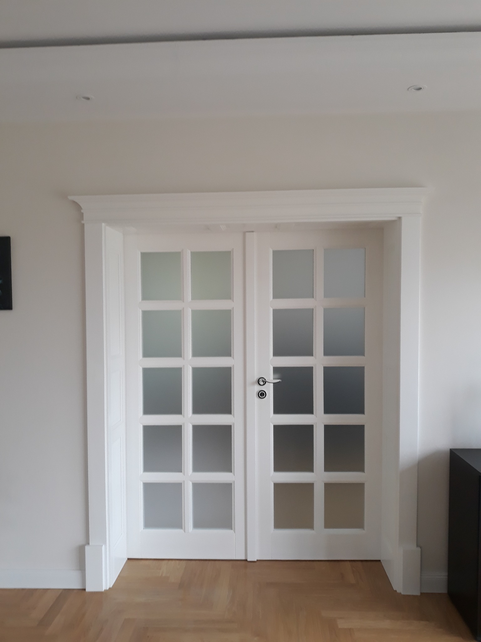 Białe, dwuskrzydłowe drzwi z matowymi szybami w ramkach, z ozdobną listwą górną, zamontowane w jasnym pomieszczeniu z parkietem.