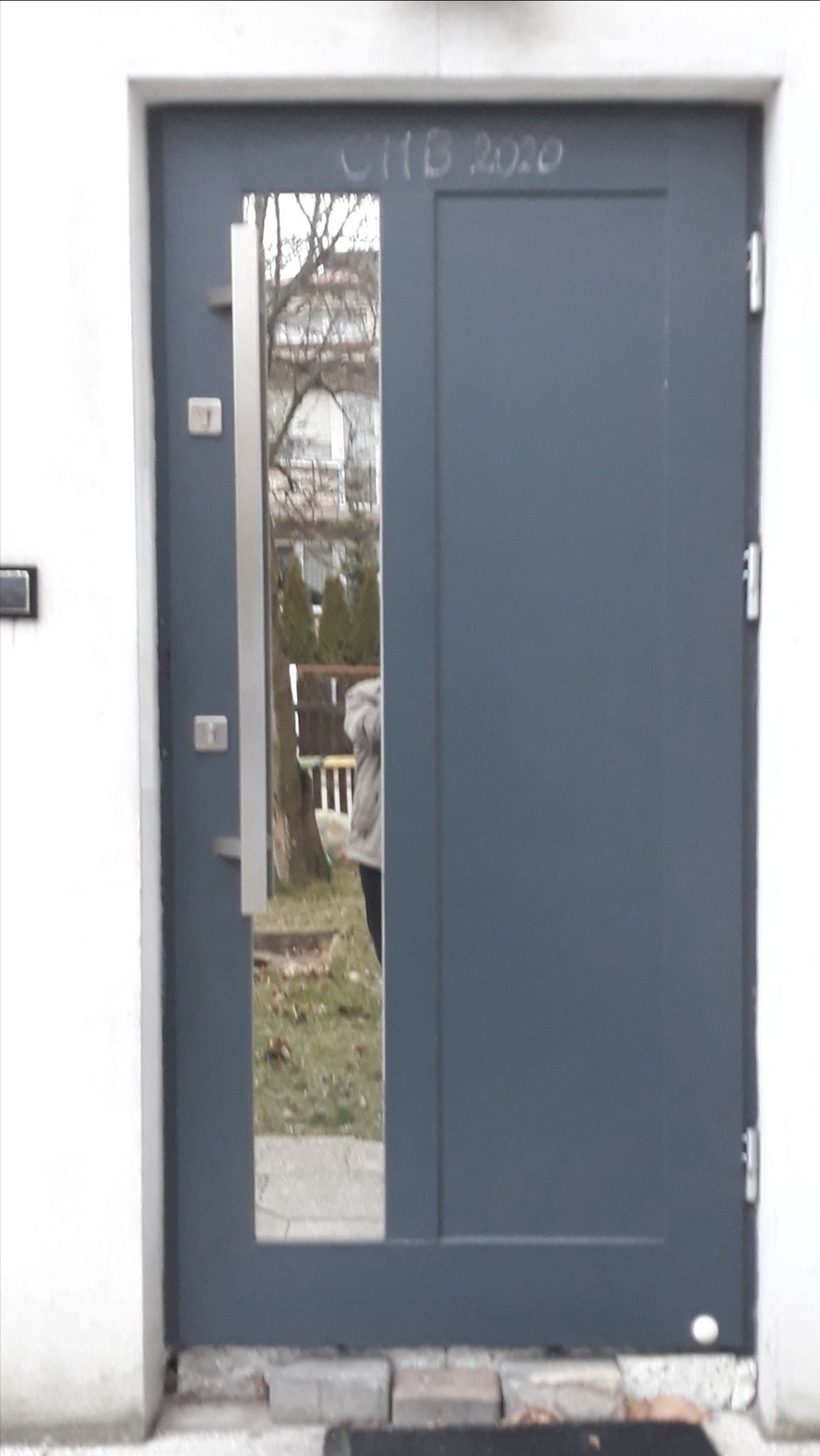 Szare drzwi wejściowe z wąskim przeszkleniem, widoczne napisy 'C11B 2020' u góry, stalowy uchwyt, odbicie osoby i ogrodu w szybie.