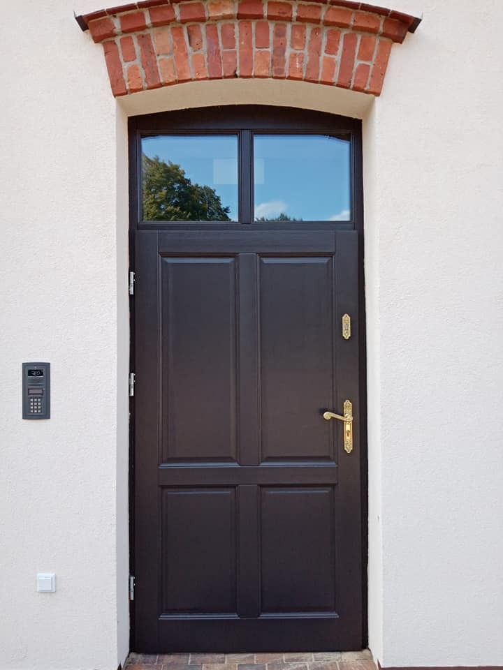 Ciemnobrązowe drzwi zewnętrzne z górnym oknem, otoczone ceglanym łukiem, zamontowane w białej ścianie z domofonem i włącznikiem światła.