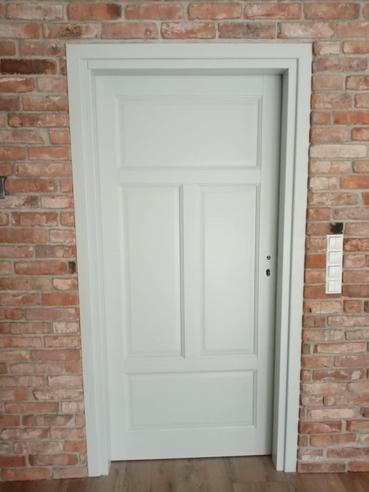 Pomalowane na jasnoszaro, trzypanelowe drzwi z widocznymi zawiasami na tle ściany z czerwonej cegły i drewnianej podłogi.
