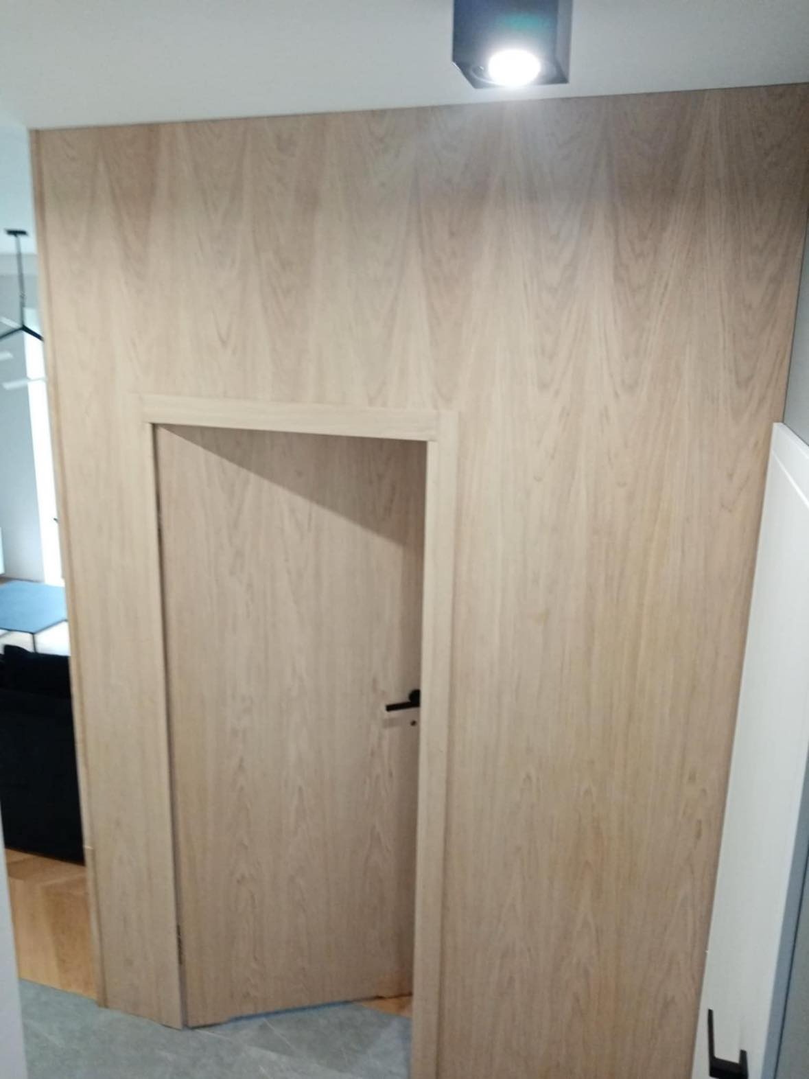 Ściana wykończona panelami drewnopodobnymi z ukrytymi drzwiami z czarną klamką, oświetlenie sufitowe LED, widok z korytarza.