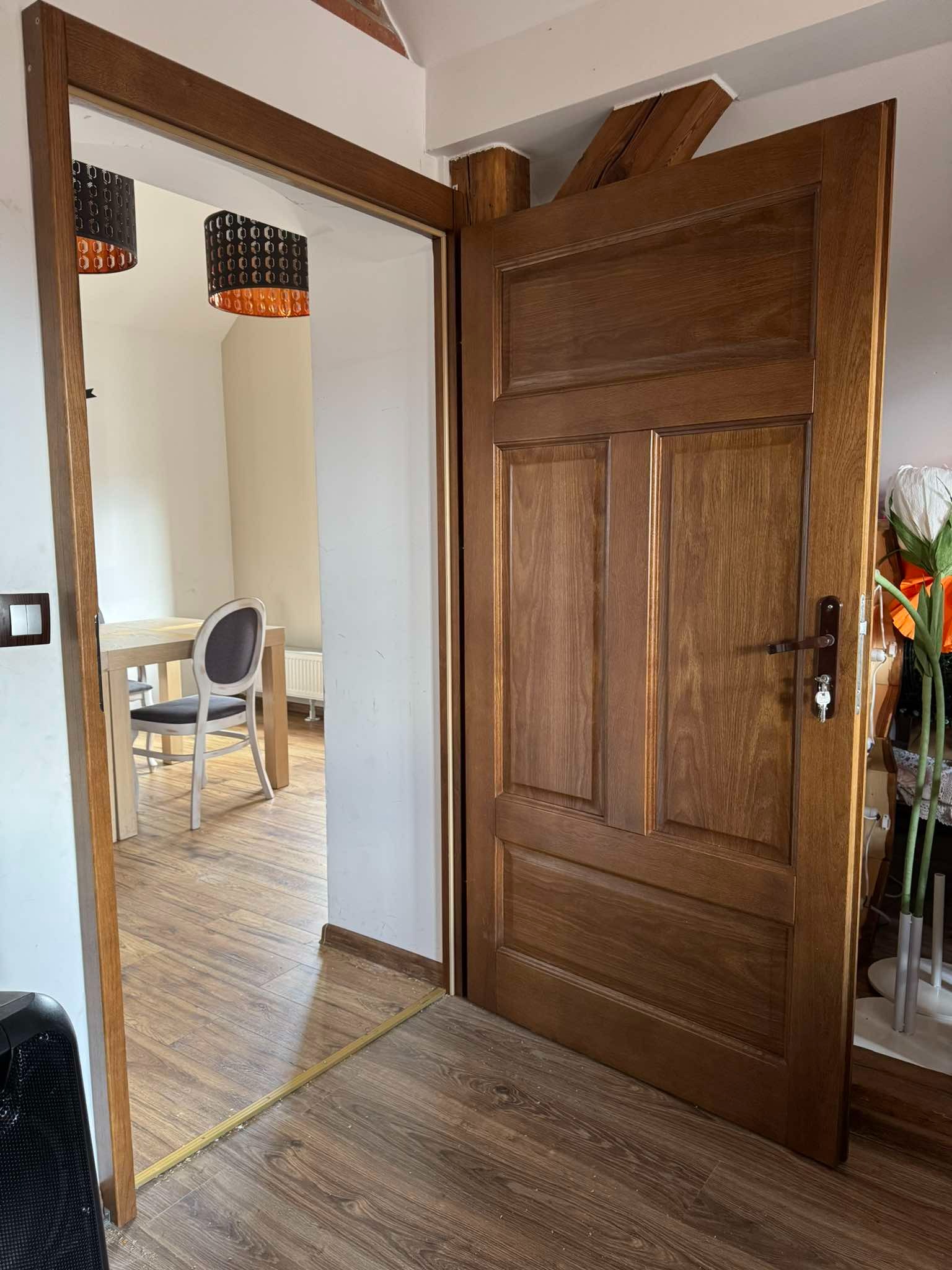 Otwarta, drewniana, trzyskrzydłowa konstrukcja w odcieniach brązu, z widocznym zamkiem i kluczem, prowadząca do jadalni z drewnianą podłogą i lampami z pomarańczowymi akcentami.