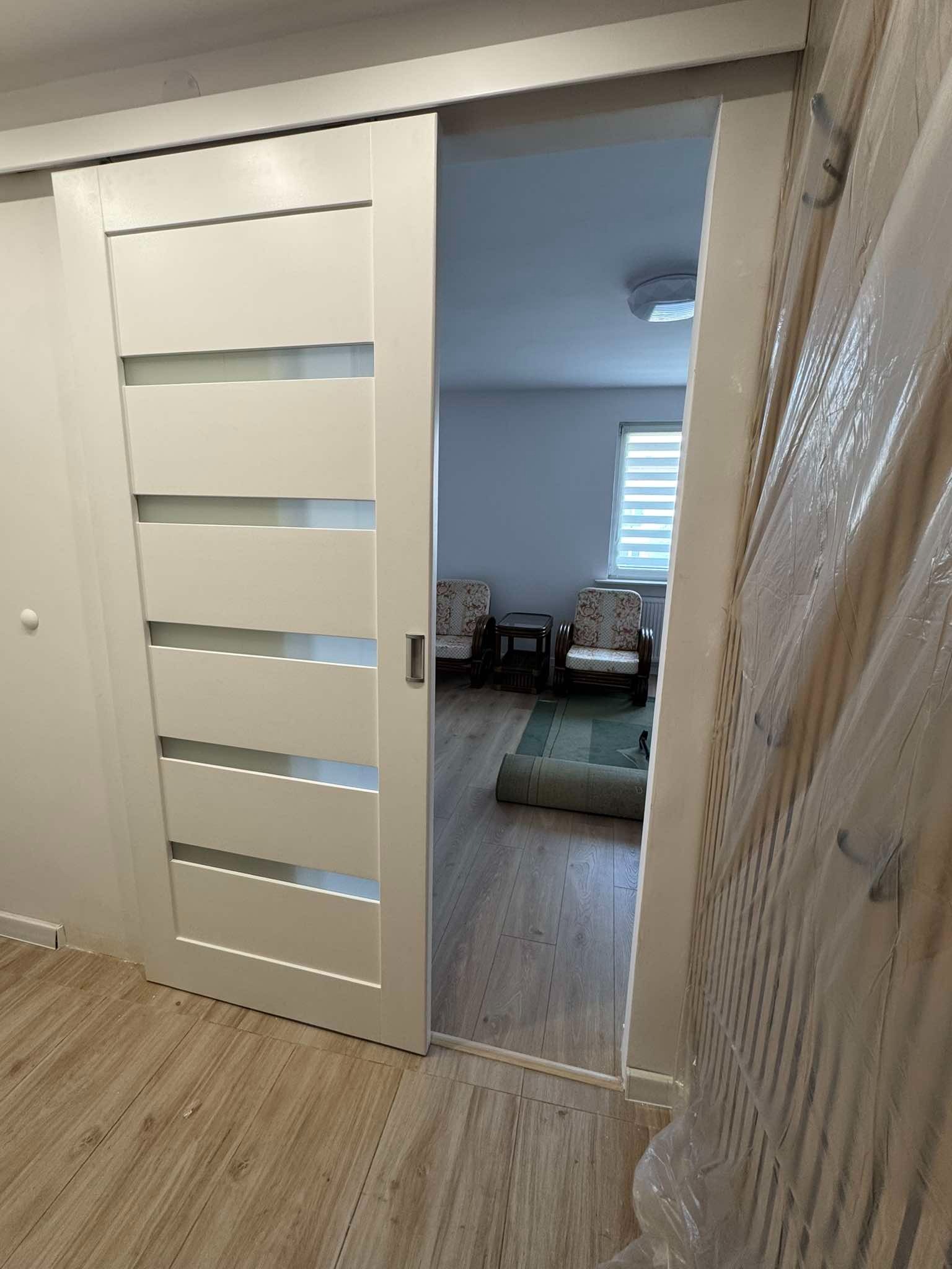 Białe drzwi przesuwne z poziomymi przeszkleniami, częściowo otwarte, widok na wnętrze pokoju z dwoma fotelami i zwiniętym dywanem, ściana obok drzwi zabezpieczona folią ochronną.