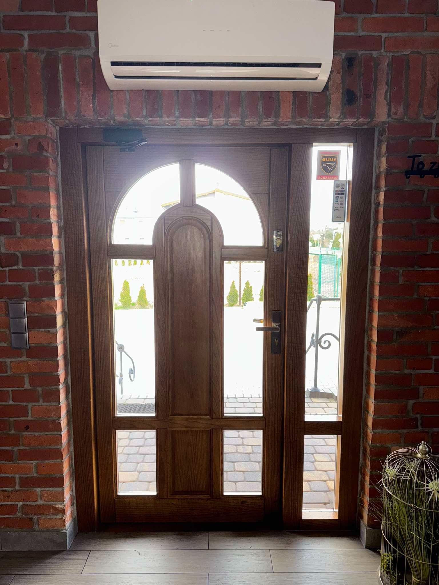 Drewniane drzwi zewnętrzne z szybami, wkomponowane w czerwoną ceglaną ścianę w Blachowni, widoczne przez szyby kostka brukowa na zewnątrz.