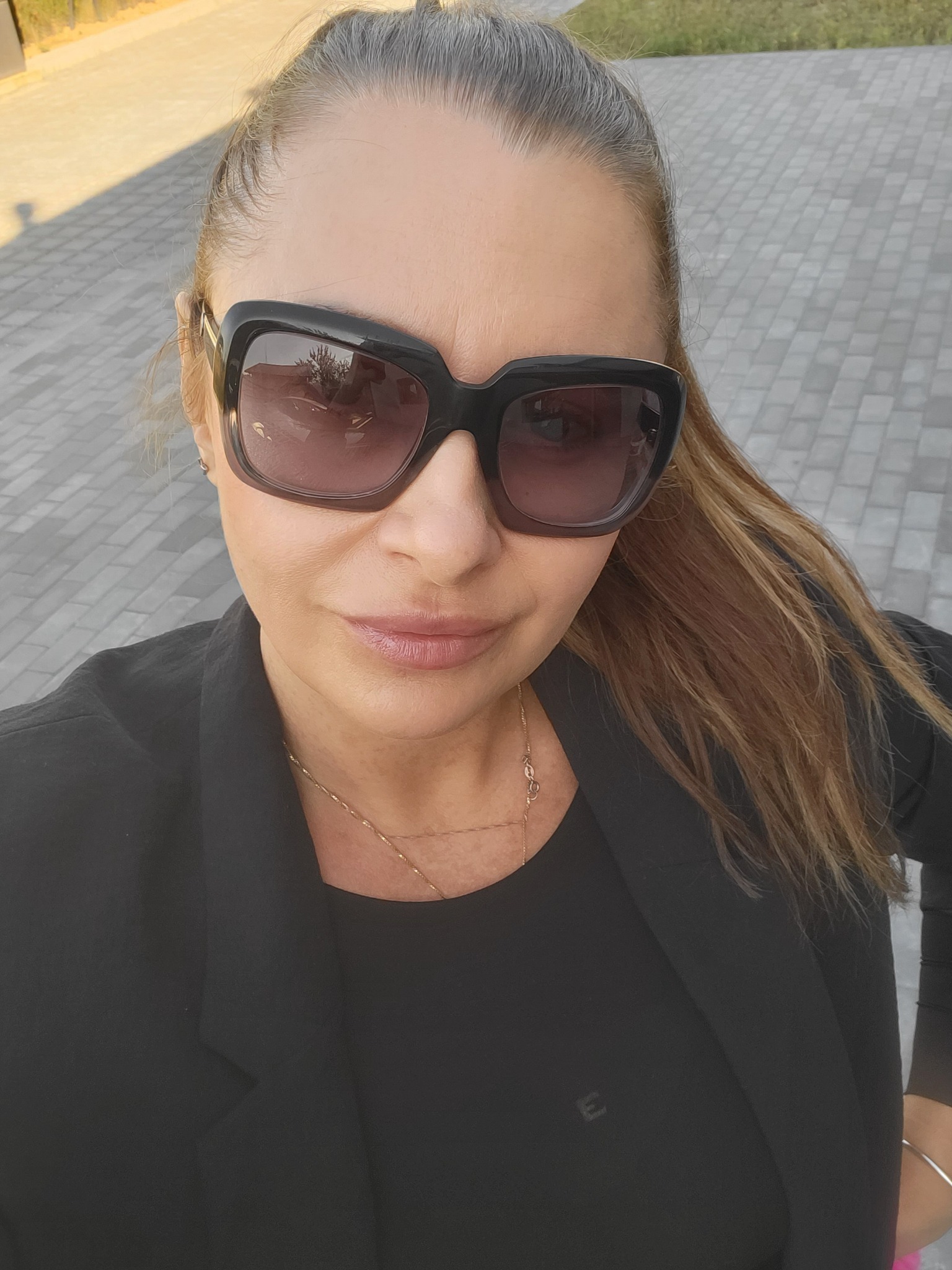 Kobieta w czarnym żakiecie i dużych okularach przeciwsłonecznych, sfotografowana z góry na tle brukowanej powierzchni.