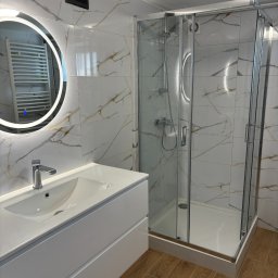 USŁUGI OGÓLNOBUDOWLANE MICHAŁ MARCINIAK - Nowoczesna łazienka po remoncie: białe marmurowe płytki, okrągłe lustro z podświetleniem LED, kabina prysznicowa ze stali i drewniana podłoga.