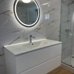 USŁUGI OGÓLNOBUDOWLANE MICHAŁ MARCINIAK - Nowoczesna łazienka z białą szafką pod umywalkę, okrągłym lustrem z podświetleniem LED i kabiną prysznicową. Marmurowe płytki na ścianach i drewnopodobna podłoga.