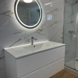 Nowoczesna łazienka z białą szafką pod umywalkę, okrągłym lustrem z podświetleniem LED i kabiną prysznicową. Marmurowe płytki na ścianach i drewnopodobna podłoga.