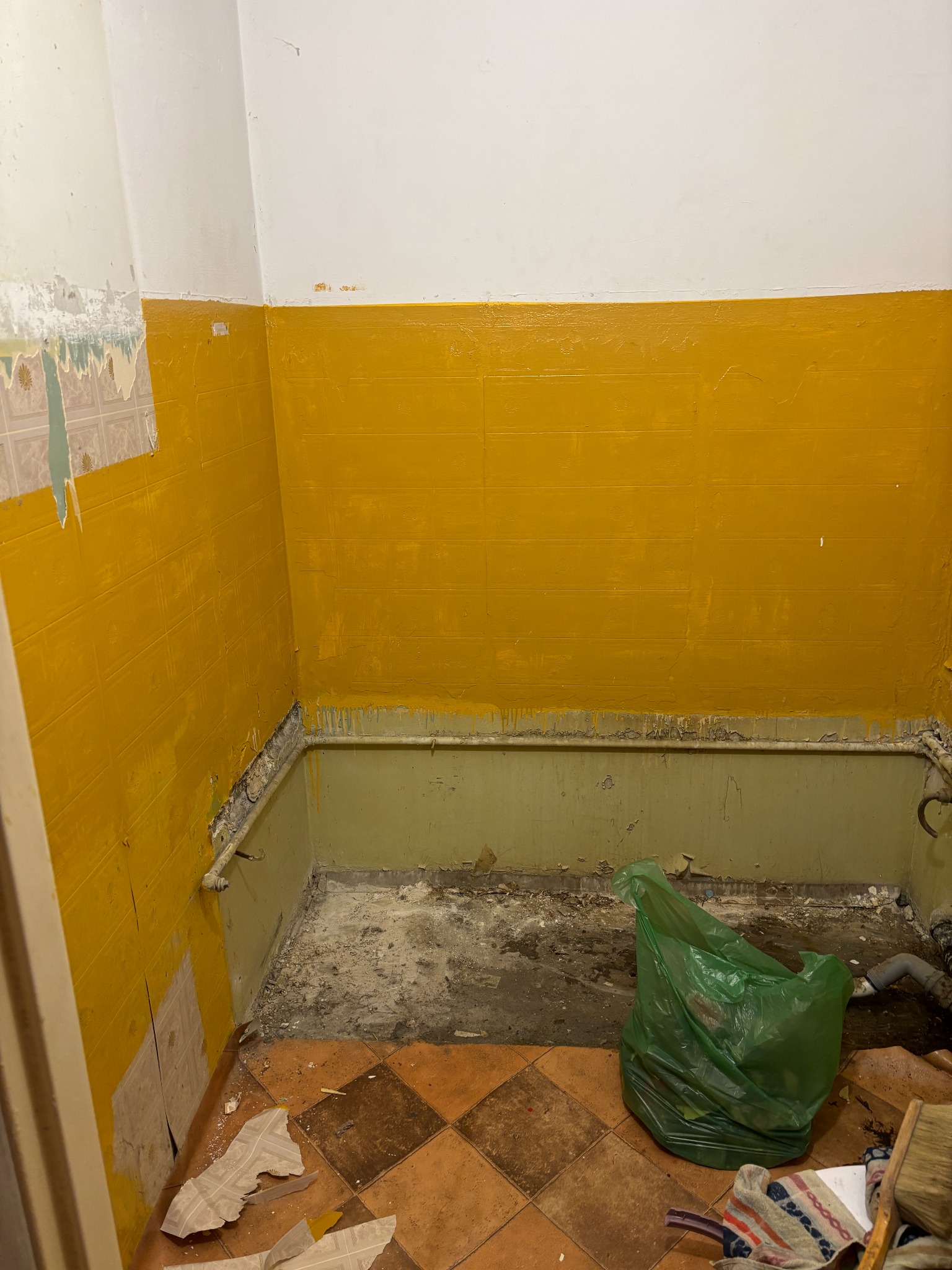 Żółte ściany w trakcie remontu łazienki, widoczne stare płytki i ślady farby, na podłodze gruz i śmieci w zielonym worku.