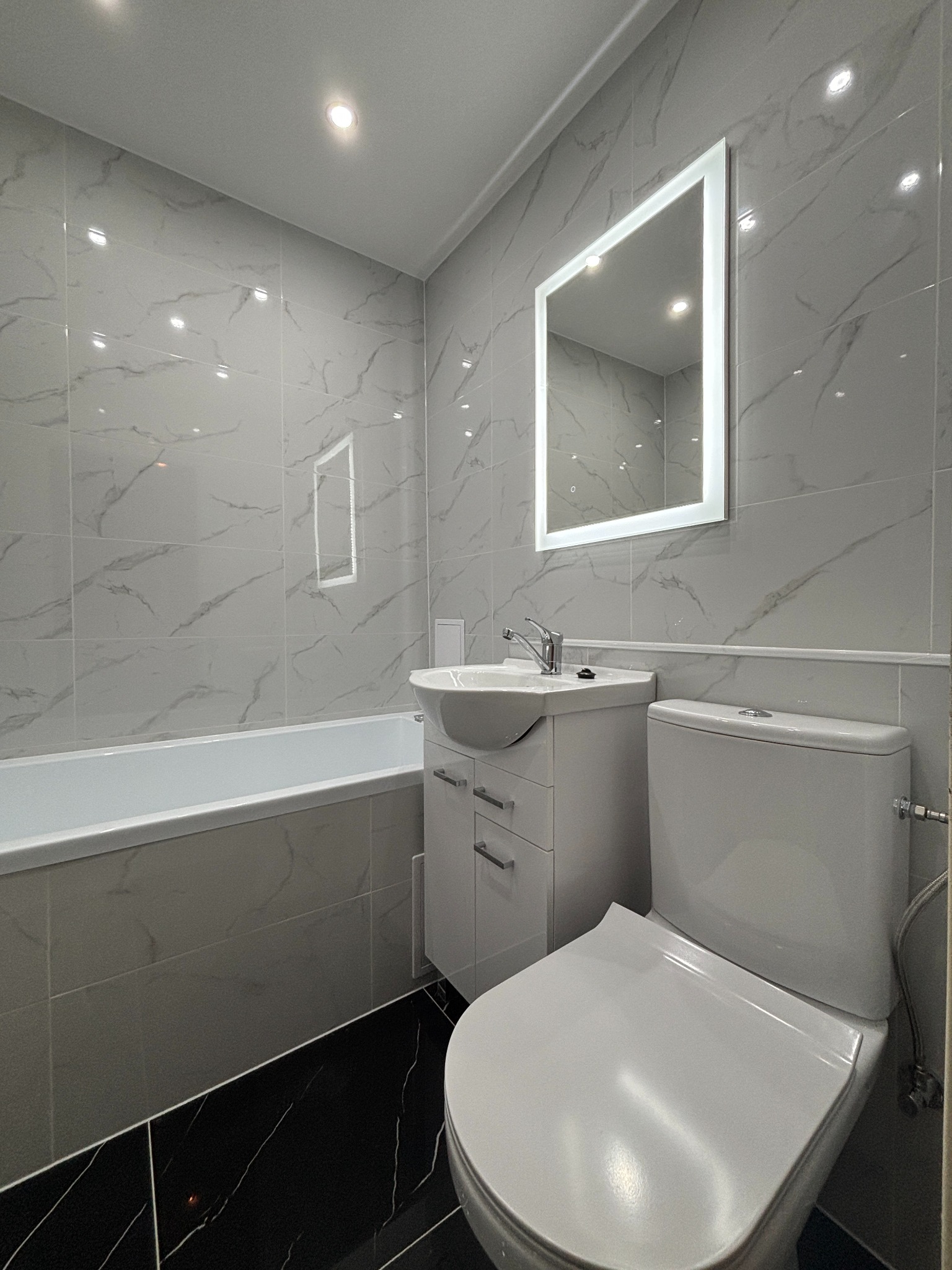 Wykończona łazienka w szaro-białej tonacji z czarną podłogą. Widoczne: wanna, umywalka z szafką, toaleta i podświetlane lustro. Ściany wyłożone płytkami imitującymi marmur.
