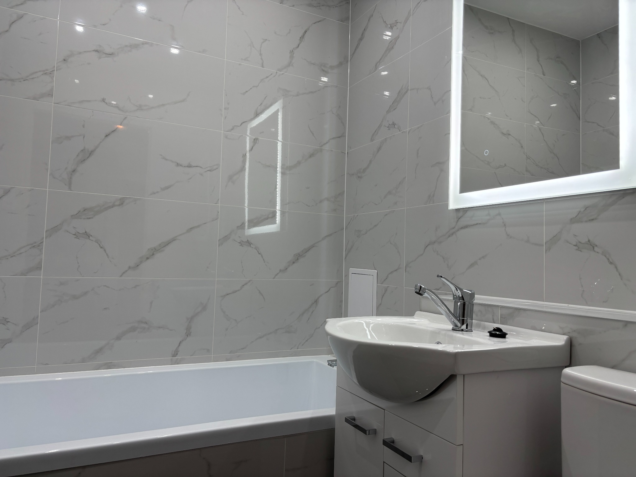 Wykończona łazienka z wanną, umywalką z szafką i podświetlanym lustrem. Ściany wyłożone płytkami imitującymi marmur w odcieniach szarości. Nowoczesny styl.