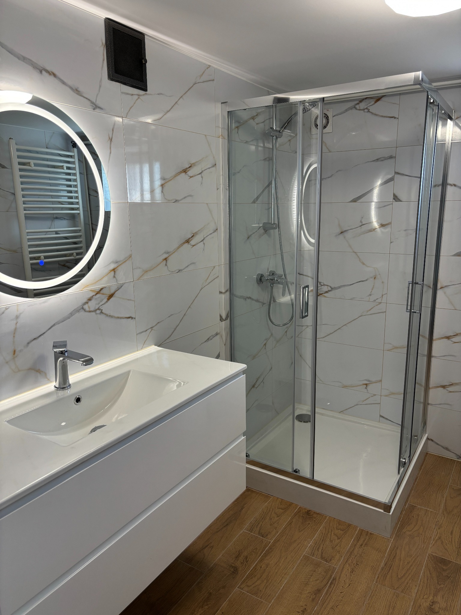 Nowoczesna łazienka po remoncie: białe marmurowe płytki, okrągłe lustro z podświetleniem LED, kabina prysznicowa ze stali i drewniana podłoga.
