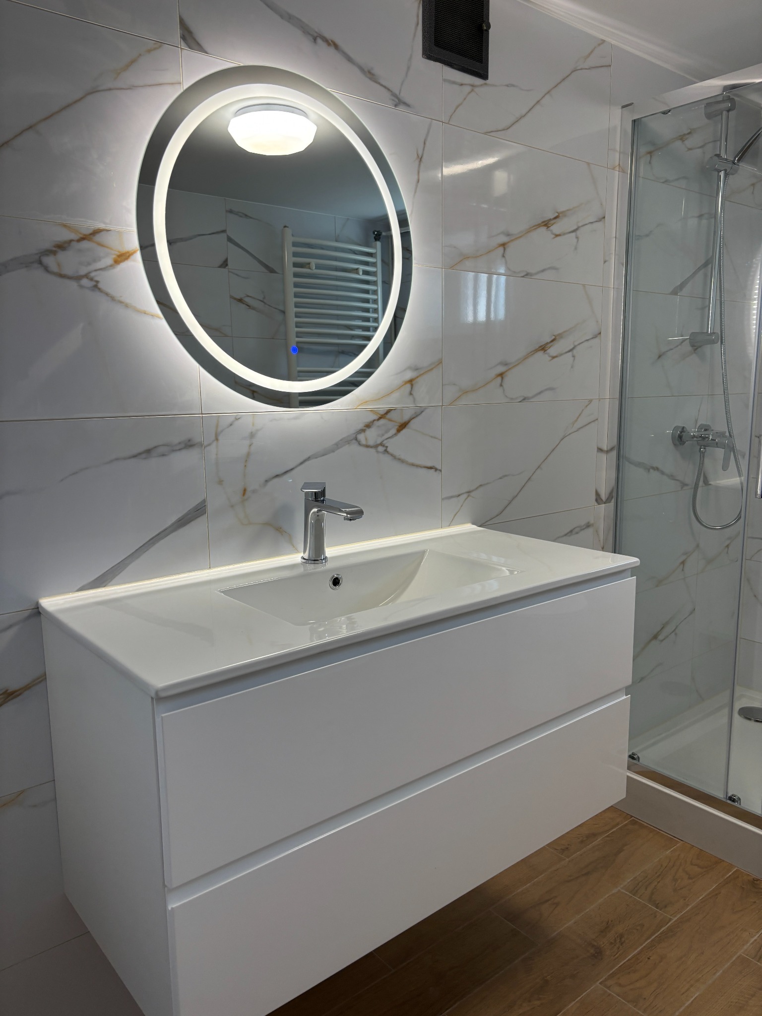 Nowoczesna łazienka z białą szafką pod umywalkę, okrągłym lustrem z podświetleniem LED i kabiną prysznicową. Marmurowe płytki na ścianach i drewnopodobna podłoga.
