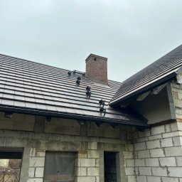 La-DACH usługi ogólnobudowlane - Dachówka ceramiczna na nowym domu w trakcie budowy, widoczny komin z cegły i system mocowań na dachu. Surowe ściany z bloczków. Pochmurne niebo w tle.