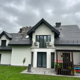 Dom z częściowo zdjętym, czarnym dachem z dachówki. Widoczne łaty i kontrłaty. Biała elewacja, szara kostka brukowa i trawnik. Pochmurne niebo w tle.