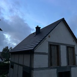 La-DACH usługi ogólnobudowlane - Dom w trakcie budowy z ciemną dachówką i kominem na tle pochmurnego nieba. Widoczne surowe ściany z pustaków i otwory okienne.