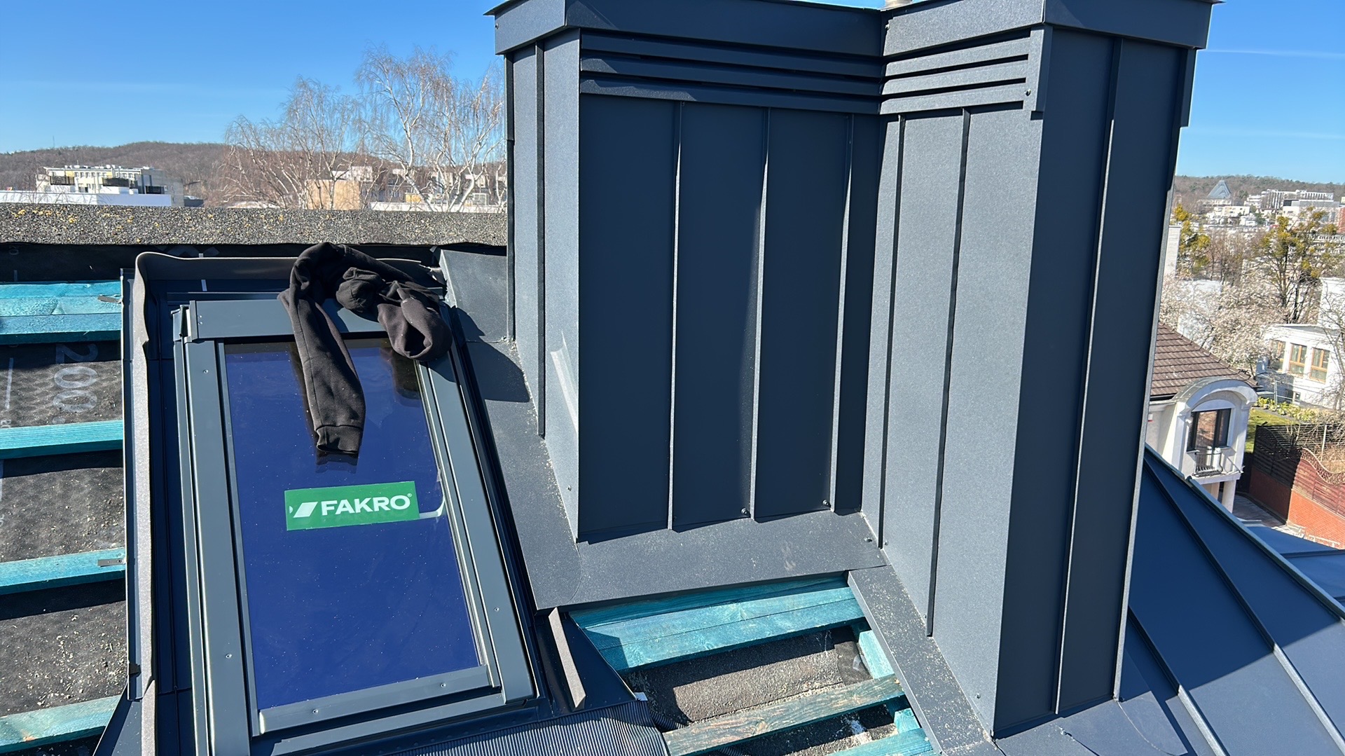 Szary komin z blachy na dachu z oknem połaciowym Fakro w słoneczny dzień. Widoczne elementy konstrukcyjne dachu i zabudowa komina z blachy na tle miejskiego krajobrazu.