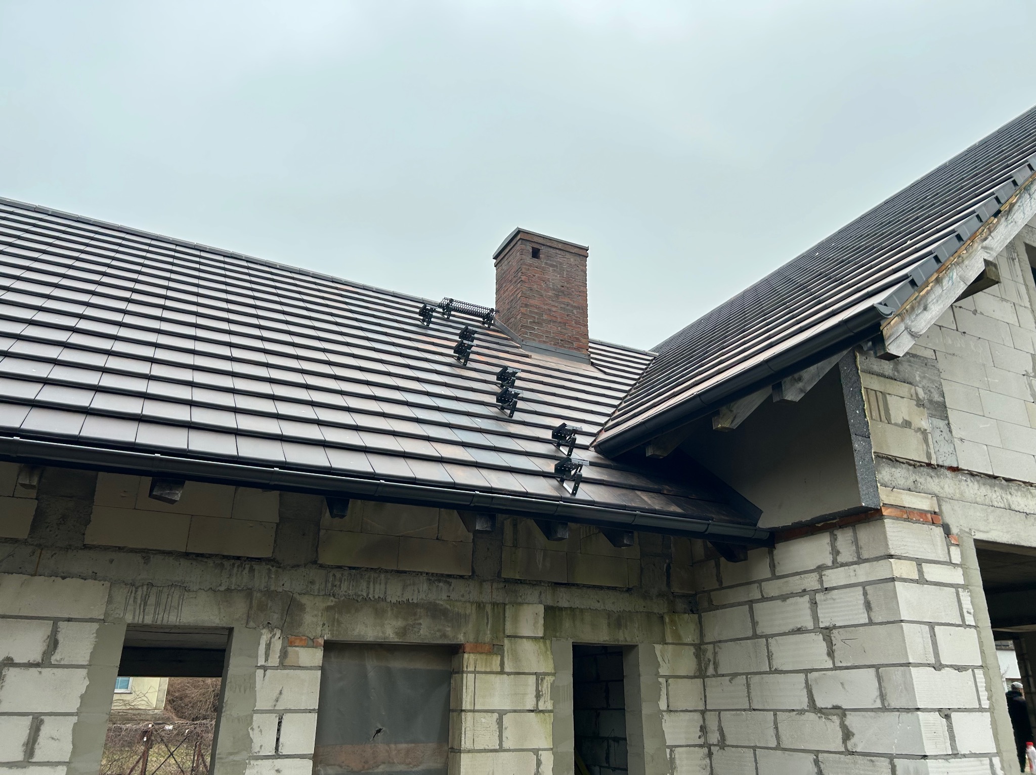 Dachówka ceramiczna na nowym domu w trakcie budowy, widoczny komin z cegły i system mocowań na dachu. Surowe ściany z bloczków. Pochmurne niebo w tle.