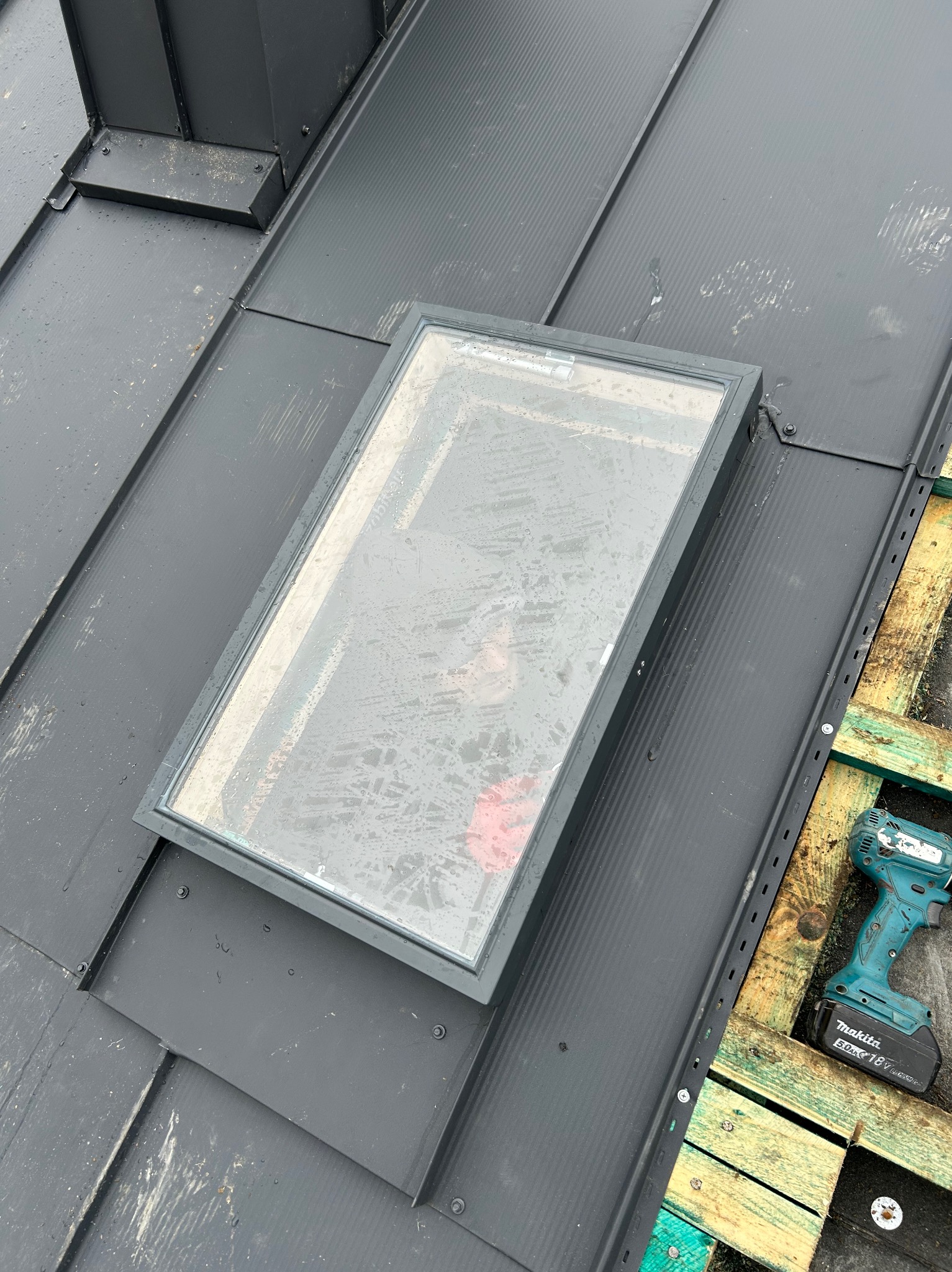 Szary dach z blachy trapezowej z zamontowanym oknem dachowym; w tle widoczny komin. W rogu wkrętarka akumulatorowa Makita na palecie.