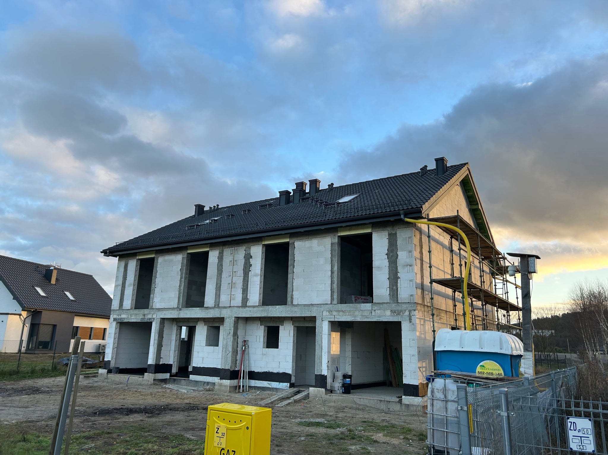 Surowy stan bliźniaka z czarnym dachem i widocznymi kominami w trakcie budowy, z żółtą skrzynką gazową i rusztowaniem z boku, na tle pochmurnego nieba.