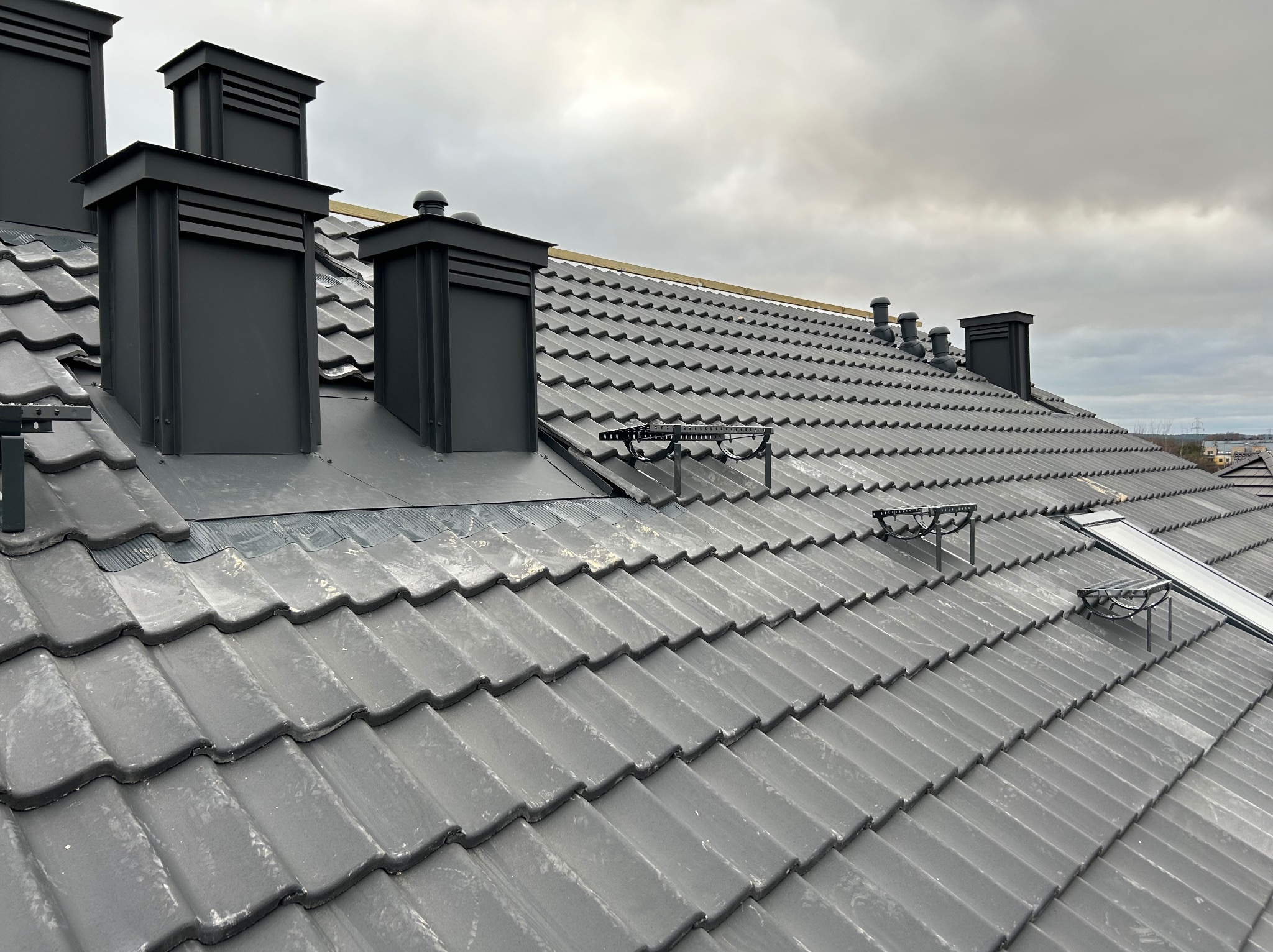 Grafitowy dach z ceramicznej dachówki karpiówki, z zamontowanymi czarnymi kominami wentylacyjnymi i zabezpieczeniami przeciwśniegowymi. Widok z góry, pochmurne niebo.