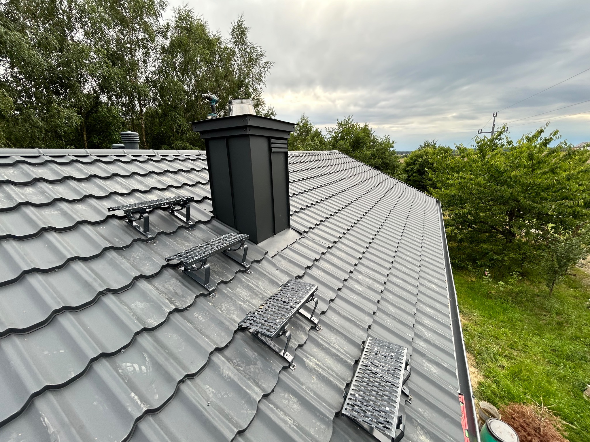 Szary dach z blachodachówki z zamontowanymi stopniami kominiarskimi i czarnym kominem na tle zieleni i pochmurnego nieba.