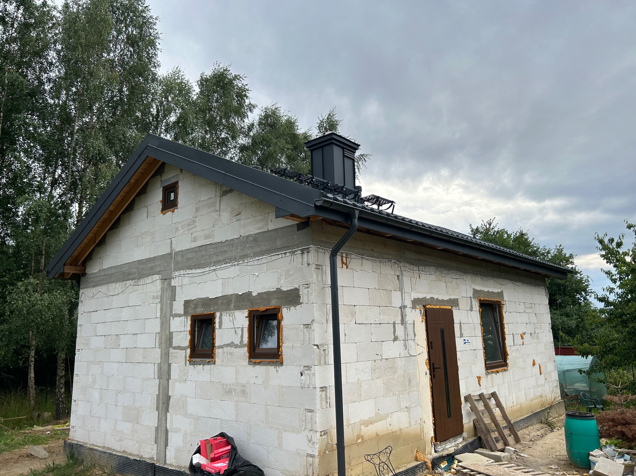 Dom w trakcie budowy, widoczne białe ściany z betonu komórkowego, okna z brązowymi ramami i czarny dach z rynnami. W tle drzewa i pochmurne niebo.
