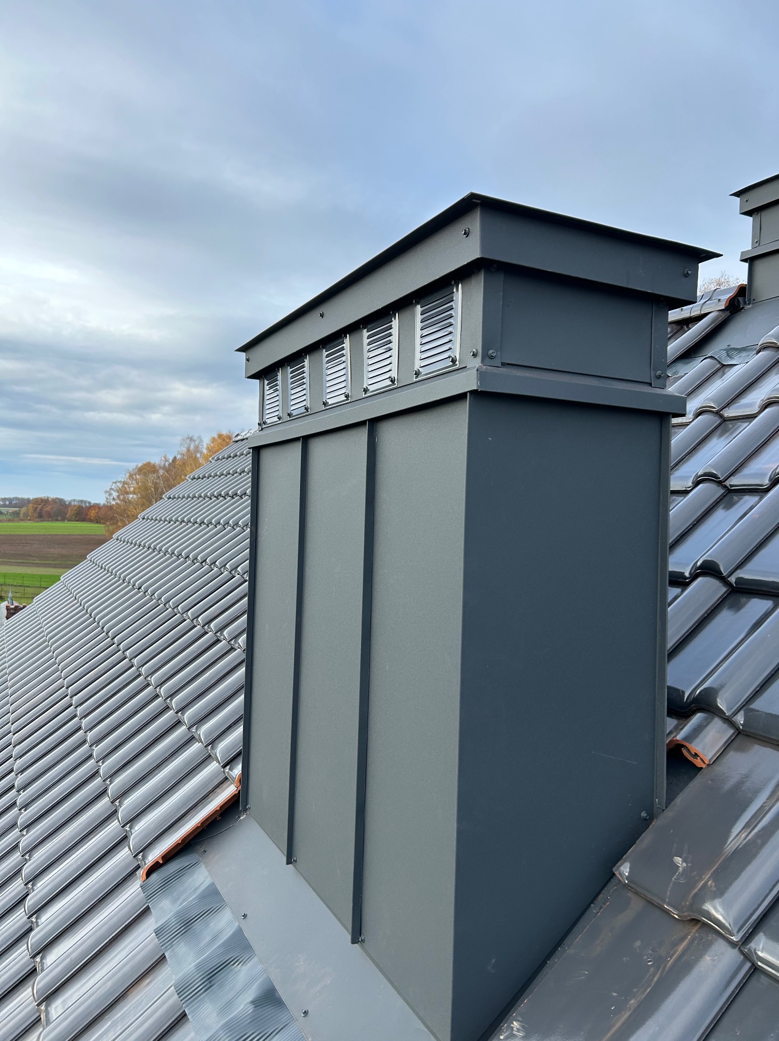 Szary komin wentylacyjny z blachy na dachu z błyszczącej dachówki ceramicznej w jesiennym krajobrazie. Widok z góry na detale wykończenia.