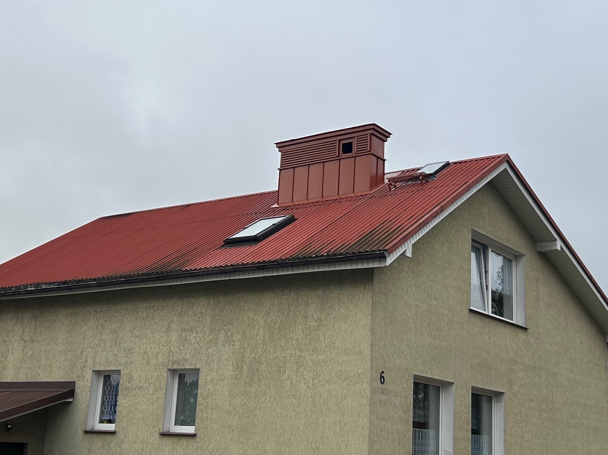 Czerwony dach z blachy falistej na budynku z beżową elewacją. Widoczne okna dachowe, komin i numer domu '6'. Pochmurne niebo w tle.