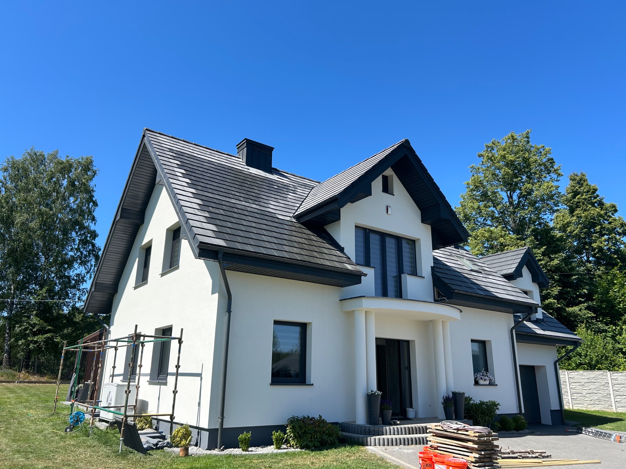 Nowoczesny dom z ciemnoszarą dachówką ceramiczną i białymi ścianami, widok z niskiej perspektywy, z rusztowaniem i materiałami budowlanymi obok.