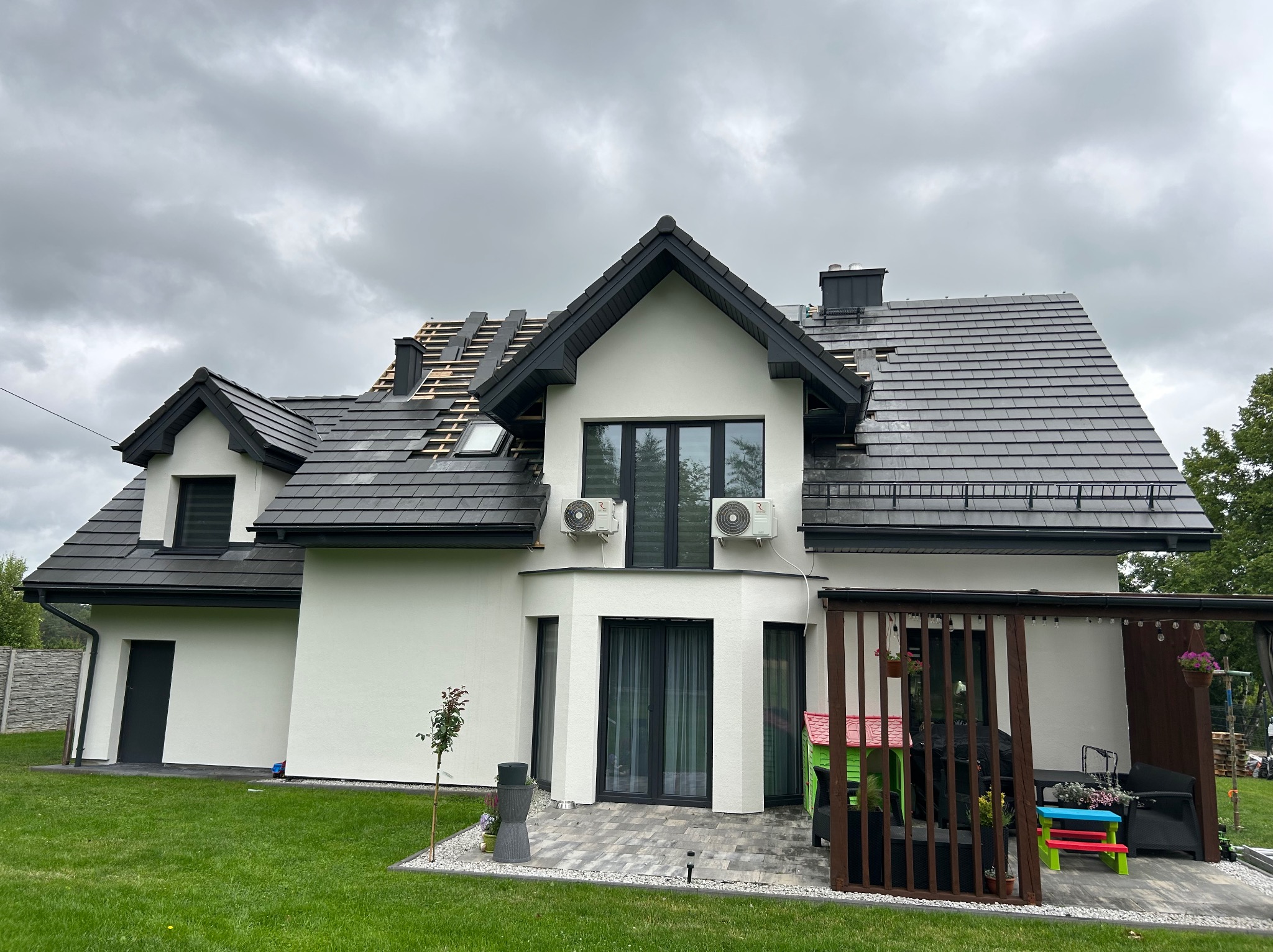 Dom z częściowo zdjętym, czarnym dachem z dachówki. Widoczne łaty i kontrłaty. Biała elewacja, szara kostka brukowa i trawnik. Pochmurne niebo w tle.