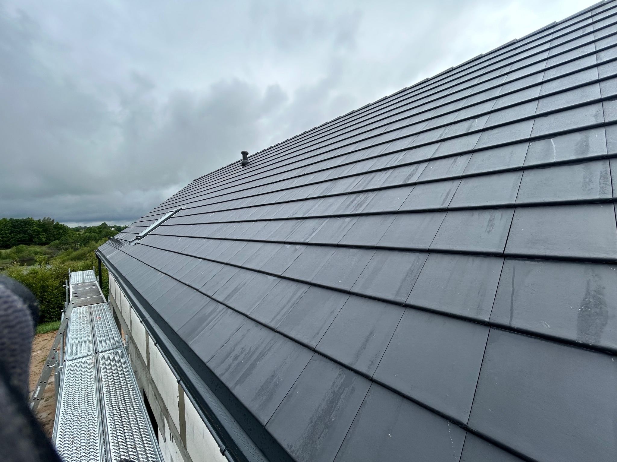 Ukończony dach z szarej dachówki ceramicznej, widok z rusztowania. Na dachu kominek wentylacyjny. Pochmurne niebo w tle.