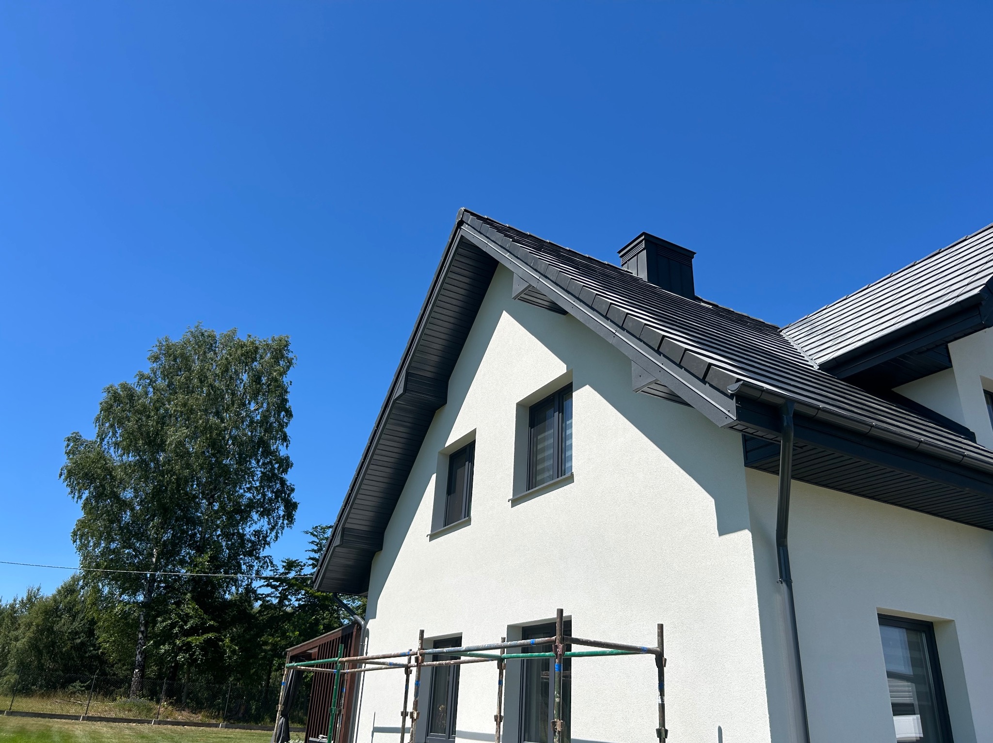 Nowoczesny, biały dom z ciemnoszarym dachem z dachówki ceramicznej i orynnowaniem. Widoczne rusztowanie przy ścianie budynku. Błękitne niebo w tle.