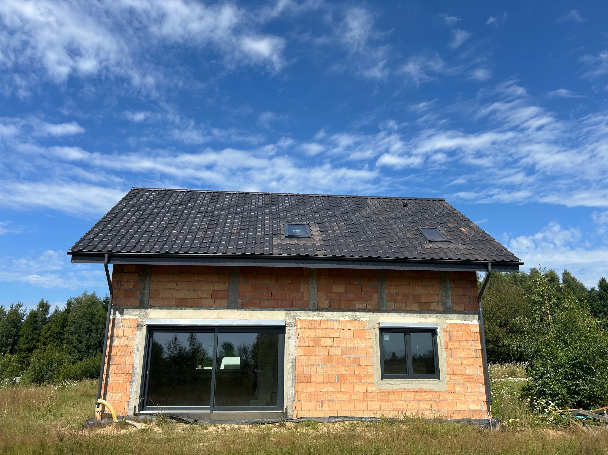 Surowy dom z ciemną dachówką na tle błękitnego nieba z chmurami. Widoczne okna i rynny. Budynek w trakcie budowy, otoczony zielenią.