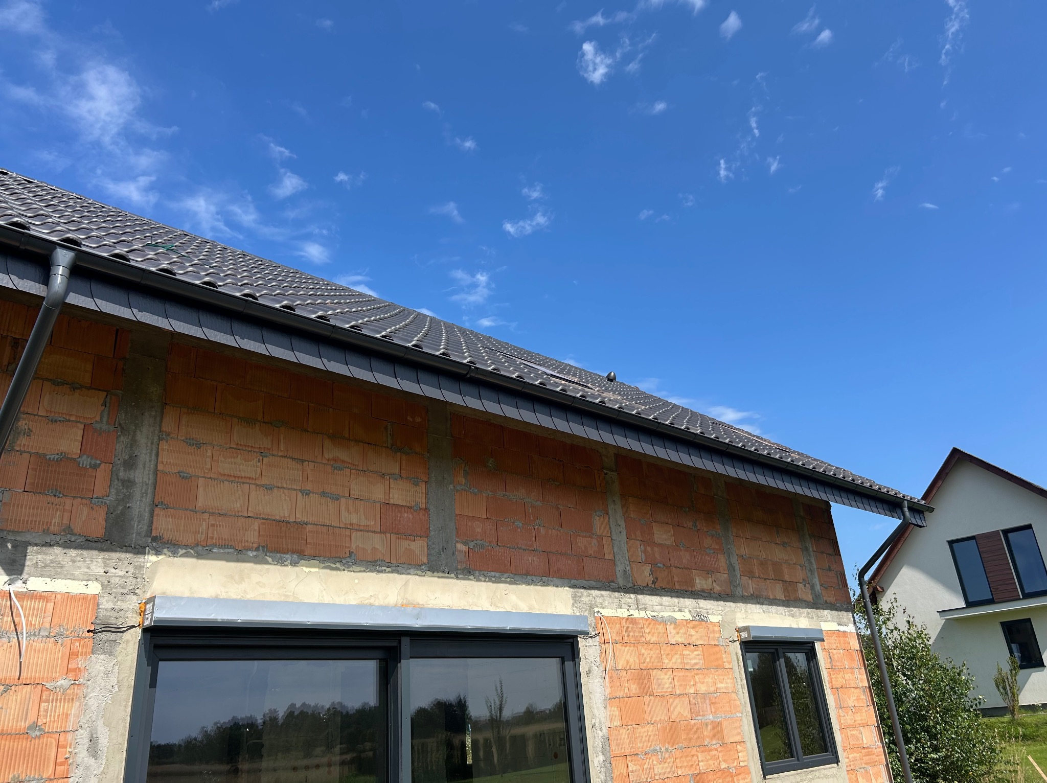 Budynek w trakcie budowy z zamontowanym dachem z ciemnej dachówki i rynnami. Widoczne okna z roletami. Błękitne niebo w tle, częściowo widoczny drugi budynek.