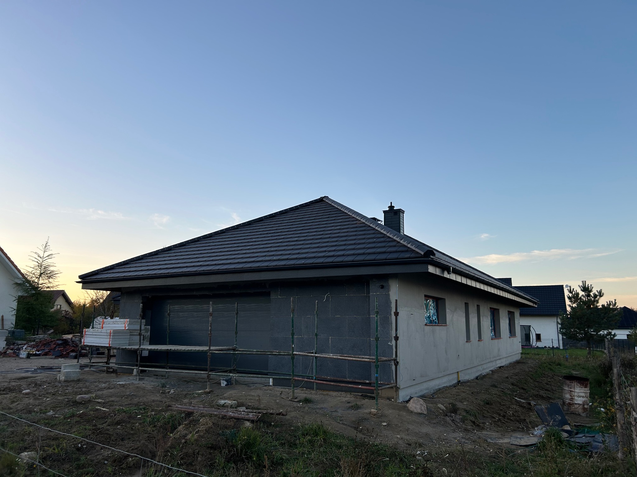 Nowoczesny dom jednorodzinny z ciemną dachówką ceramiczną, w trakcie prac wykończeniowych elewacji. Widoczne rusztowania i materiały budowlane.