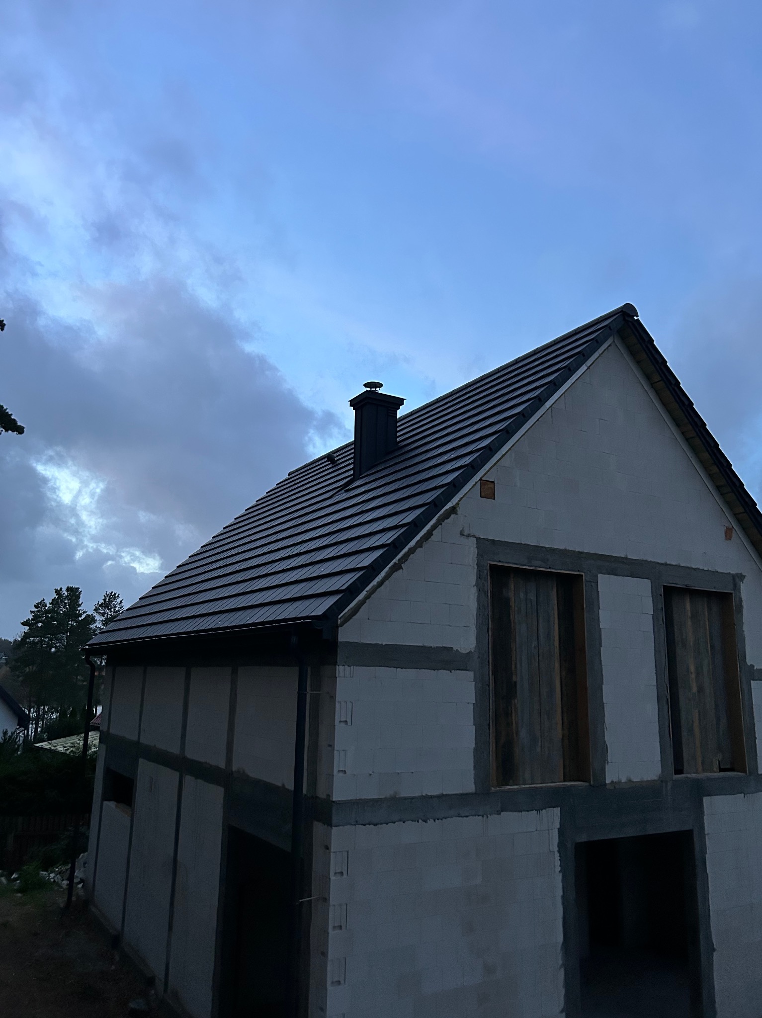Dom w trakcie budowy z ciemną dachówką i kominem na tle pochmurnego nieba. Widoczne surowe ściany z pustaków i otwory okienne.