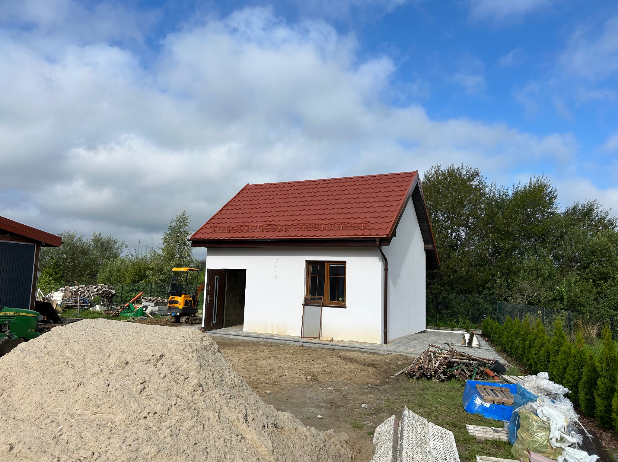 Biały budynek z czerwoną dachówką w trakcie budowy, widoczny stos piasku i sprzęt budowlany. Częściowo ułożona kostka brukowa.