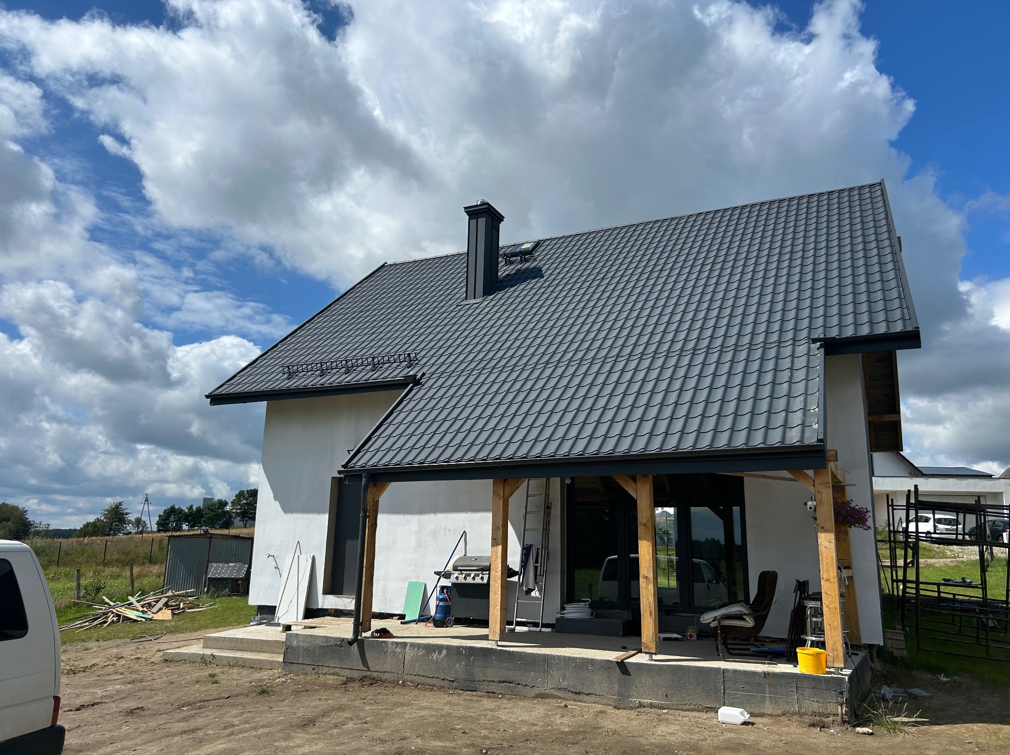 Dom jednorodzinny z szarą dachówką modułową, widok z zewnątrz w słoneczny dzień. Na dachu widoczny komin i okno dachowe. Częściowo widoczny samochód dostawczy.