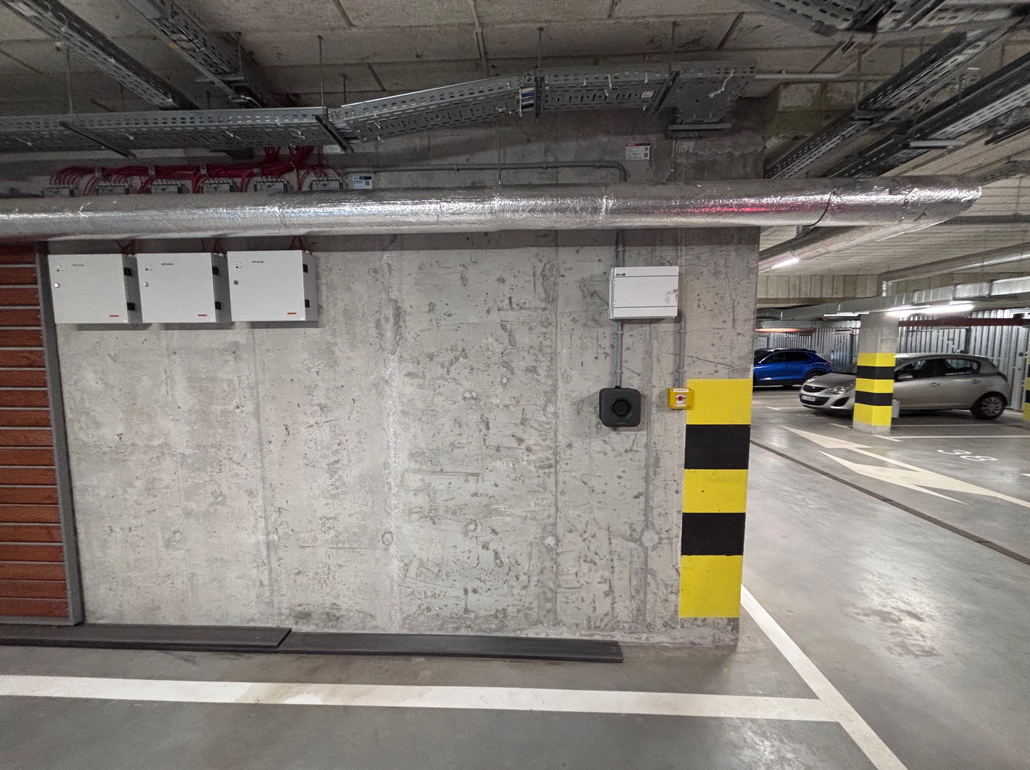 Szare skrzynki elektryczne zamontowane na betonowej ścianie w garażu podziemnym, widoczne kable w korytach pod sufitem.