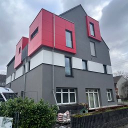 EURO-THERM SPÓŁKA Z OGRANICZONĄ ODPOWIEDZIALNOŚCIĄ - Nowoczesny budynek z szarą elewacją i czerwonymi, kubistycznymi elementami okiennymi, widok z zewnątrz na tle pochmurnego nieba, worki z materiałami budowlanymi przy ogrodzeniu.