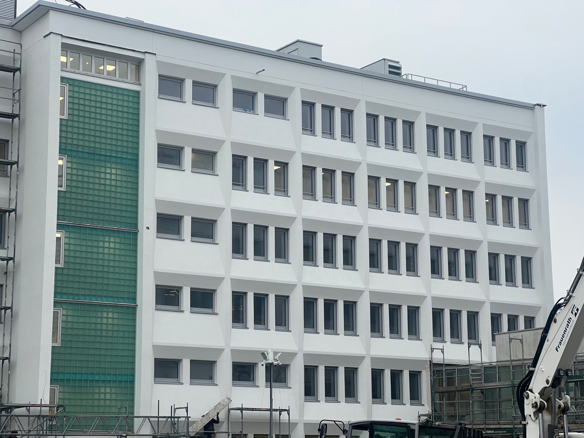 Wielopiętrowy budynek biurowy z białą elewacją, widoczne rusztowania i fragment dźwigu podczas prac budowlanych.