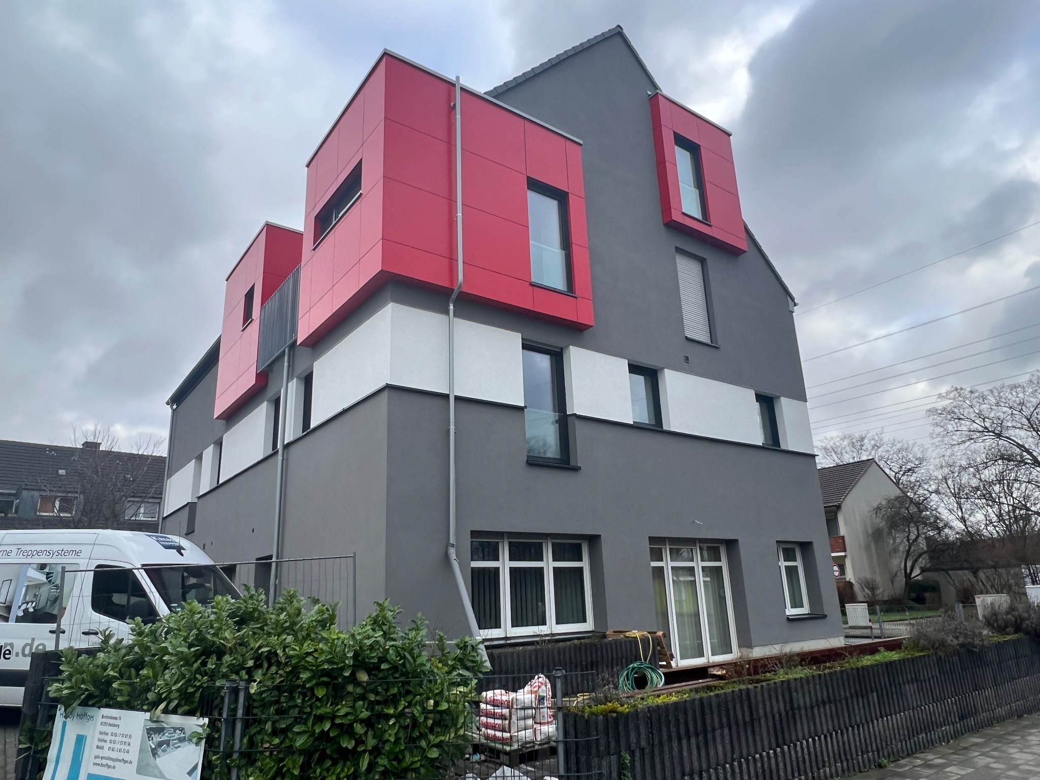 Nowoczesny budynek z szarą elewacją i czerwonymi, kubistycznymi elementami okiennymi, widok z zewnątrz na tle pochmurnego nieba, worki z materiałami budowlanymi przy ogrodzeniu.