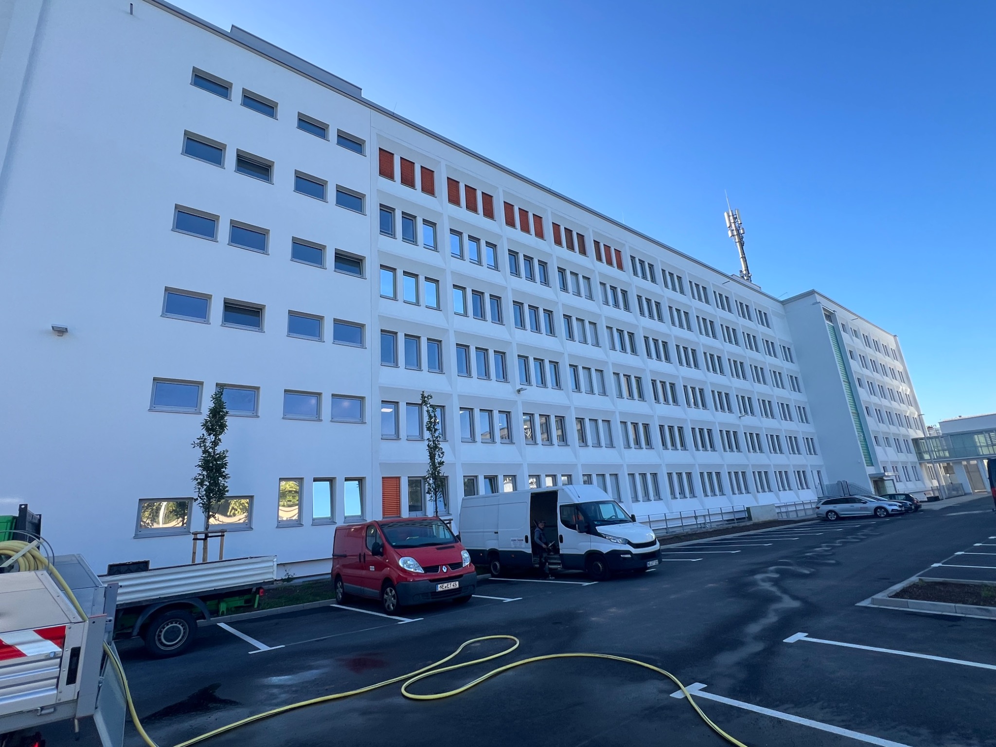 Ocieplony budynek biurowy z elewacją, widoczne samochody dostawcze i drzewa. Część okien z pomarańczowymi żaluzjami. Nowe, czarne parkingi. Wąż na pierwszym planie.