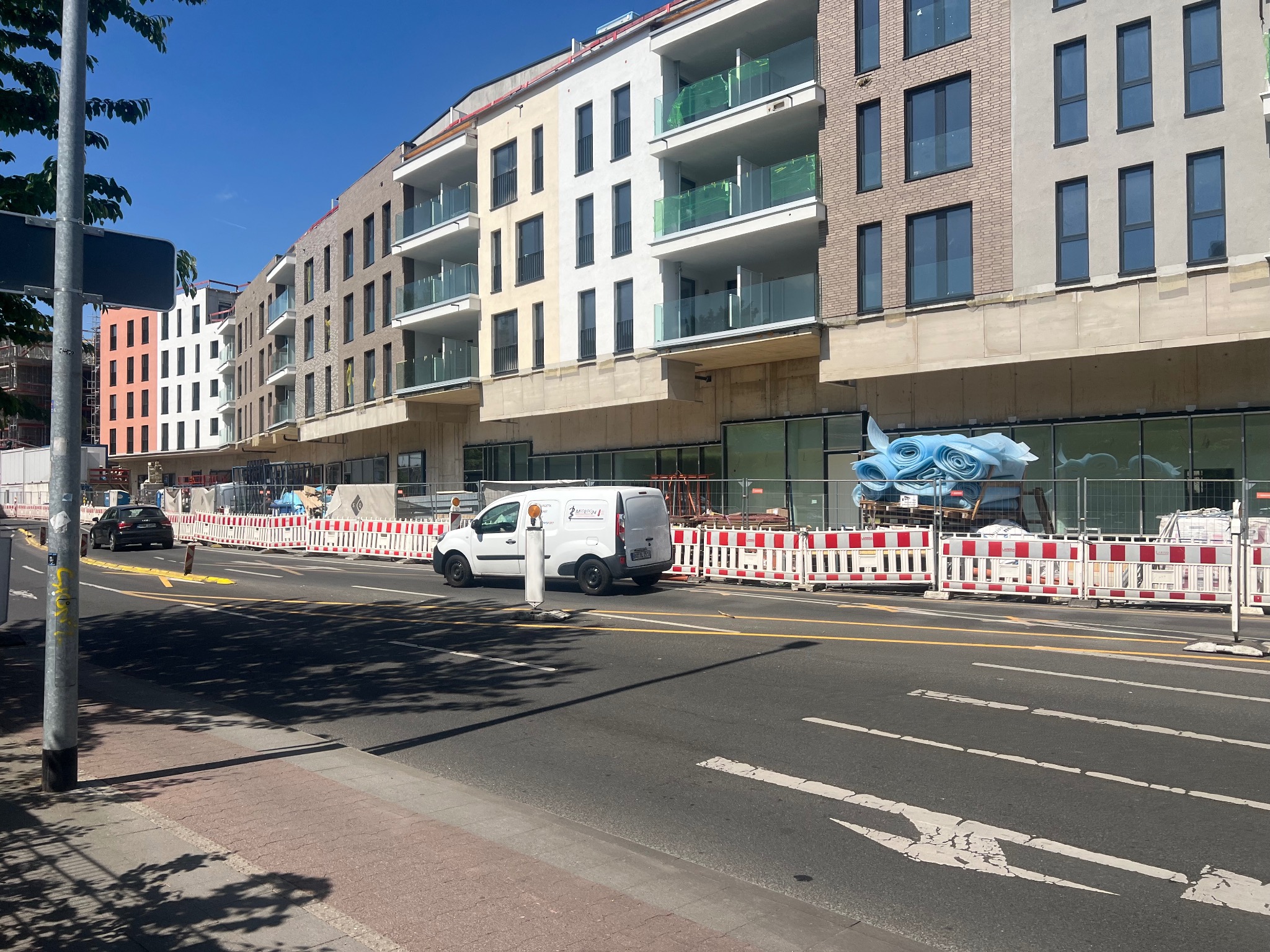 Wielopiętrowy budynek w trakcie budowy/wykończenia z widoczną elewacją w różnych odcieniach beżu, bieli i brązu. Na pierwszym planie barierki i samochody.