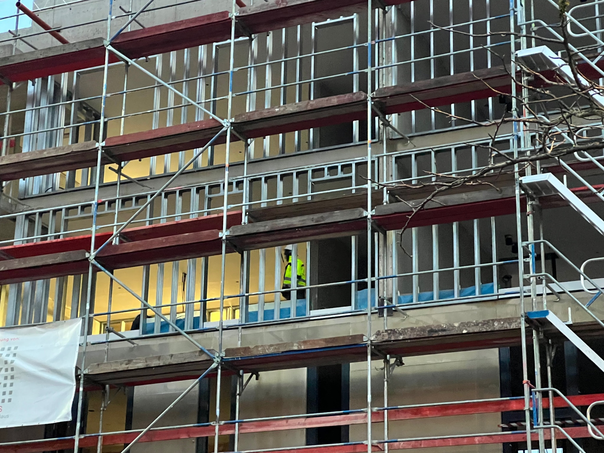 Budynek w trakcie ocieplania, widoczny przez rusztowanie. Pracownik w żółtej kamizelce w oknie. Elementy elewacji przygotowane do montażu.