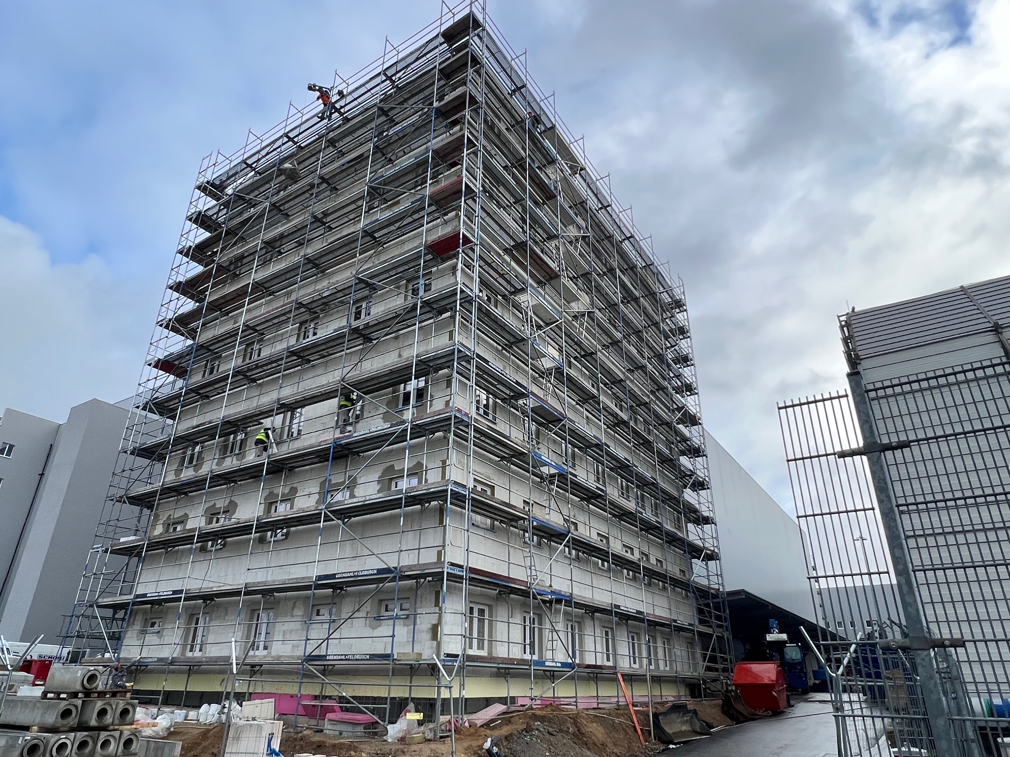 Wielopiętrowy budynek w trakcie prac elewacyjnych, otoczony rusztowaniami. Widoczni pracownicy na różnych poziomach. Ujęcie z dołu, pochmurne niebo w tle.